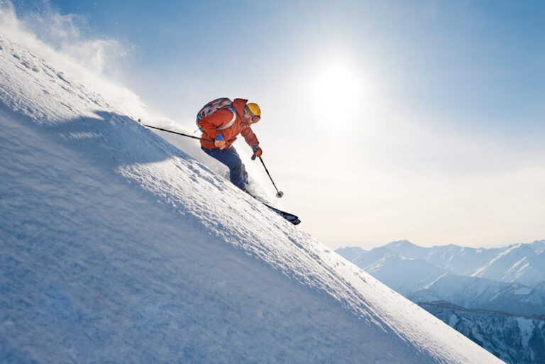 Skifahrer in roter Jacke fährt steil einen verschneiten Hang hinab, Sonne und Berggipfel im Hintergrund