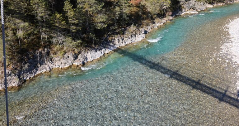 Klares, flaches Bergflusswasser mit sichtbarem kiesigem Grund, felsig-bewaldetes Ufer, kleiner Holzsteg und diagonaler Brücke