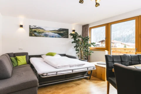 Appartementzimmer mit Bett, Holzfu&szlig;boden, gro&szlig;em Fenster mit Bergblick, Esstisch, Zimmerpflanze und Wandbild.
