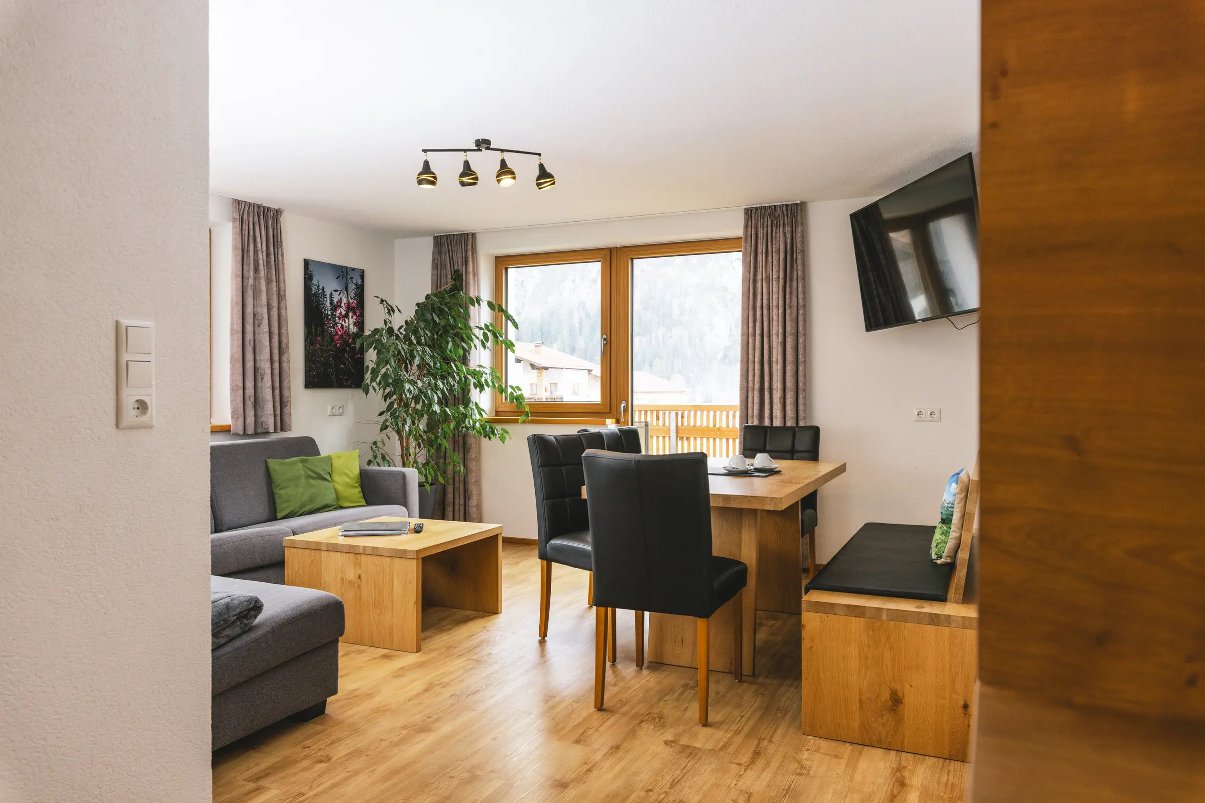 Wohnzimmer einer Ferienwohnung mit Holzboden, Eckbank und Esstisch, Sofa, Zimmerpflanze, Wand-TV und gro&szlig;em Fenster
