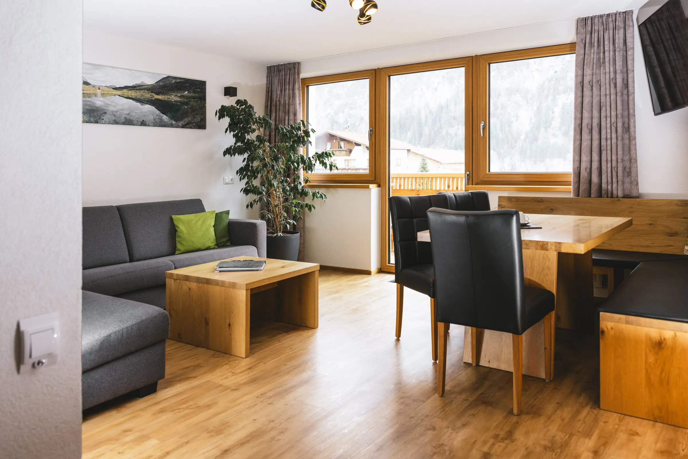 Wohn- und Essbereich einer Ferienwohnung in Holzgau mit grauem Sofa, Holzboden, Esstisch, gro&szlig;en Fenstern und Bergpanorama
